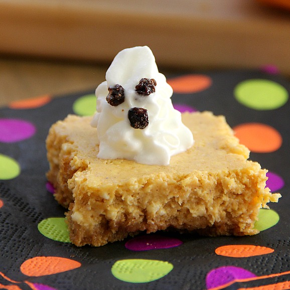Pumpkin Cheesecake Bars on a wooden board with whipped cream ghosts on top on a polka dot napkin