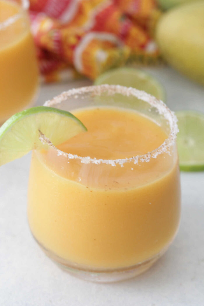 Angled photo of a mango margarita in a class with sliced lime wedges and brightly colored cloth napkin in the background