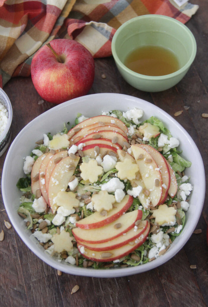 Angled photo of brussels sprouts salad in a white bowl with sliced apples and crumbled goat cheese and a red and orange napkin