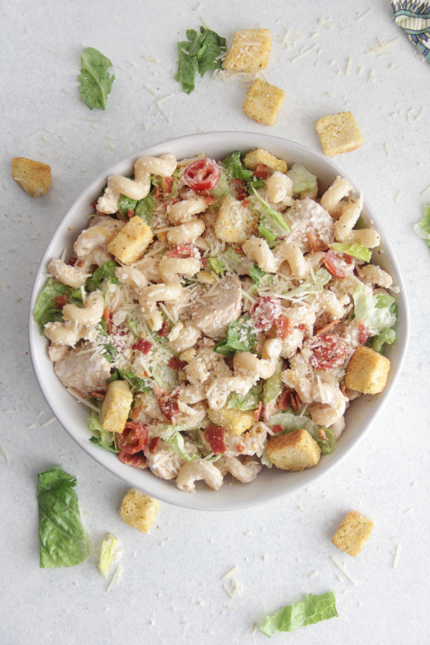 Overhead of a bowl of Chicken Caesar Pasta Salad in a white bowl with croutons and lettuce scattered around the side
