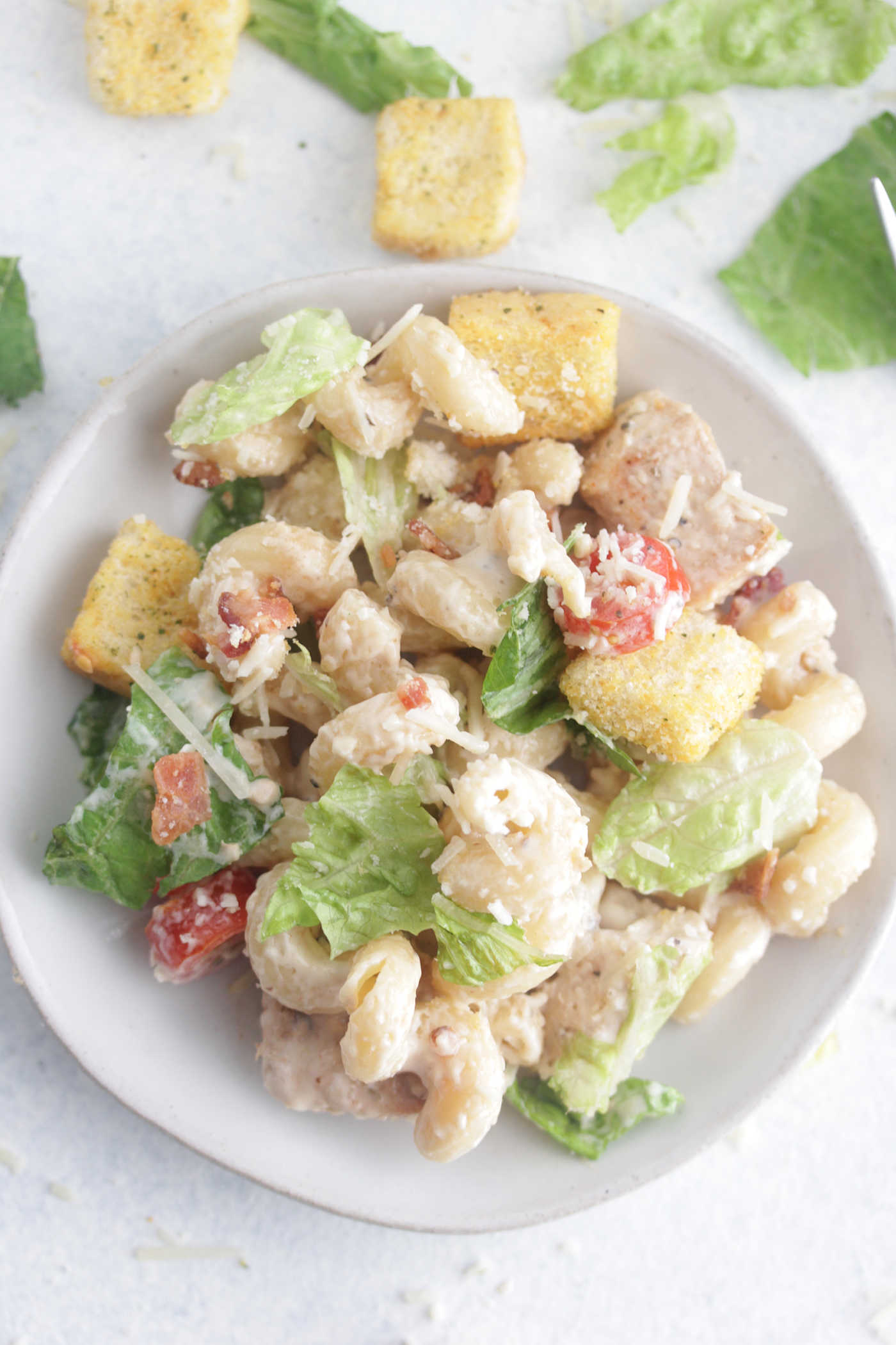 Chicken Caesar Pasta Salad on a small white plate with scattered parmesan cheese around the side