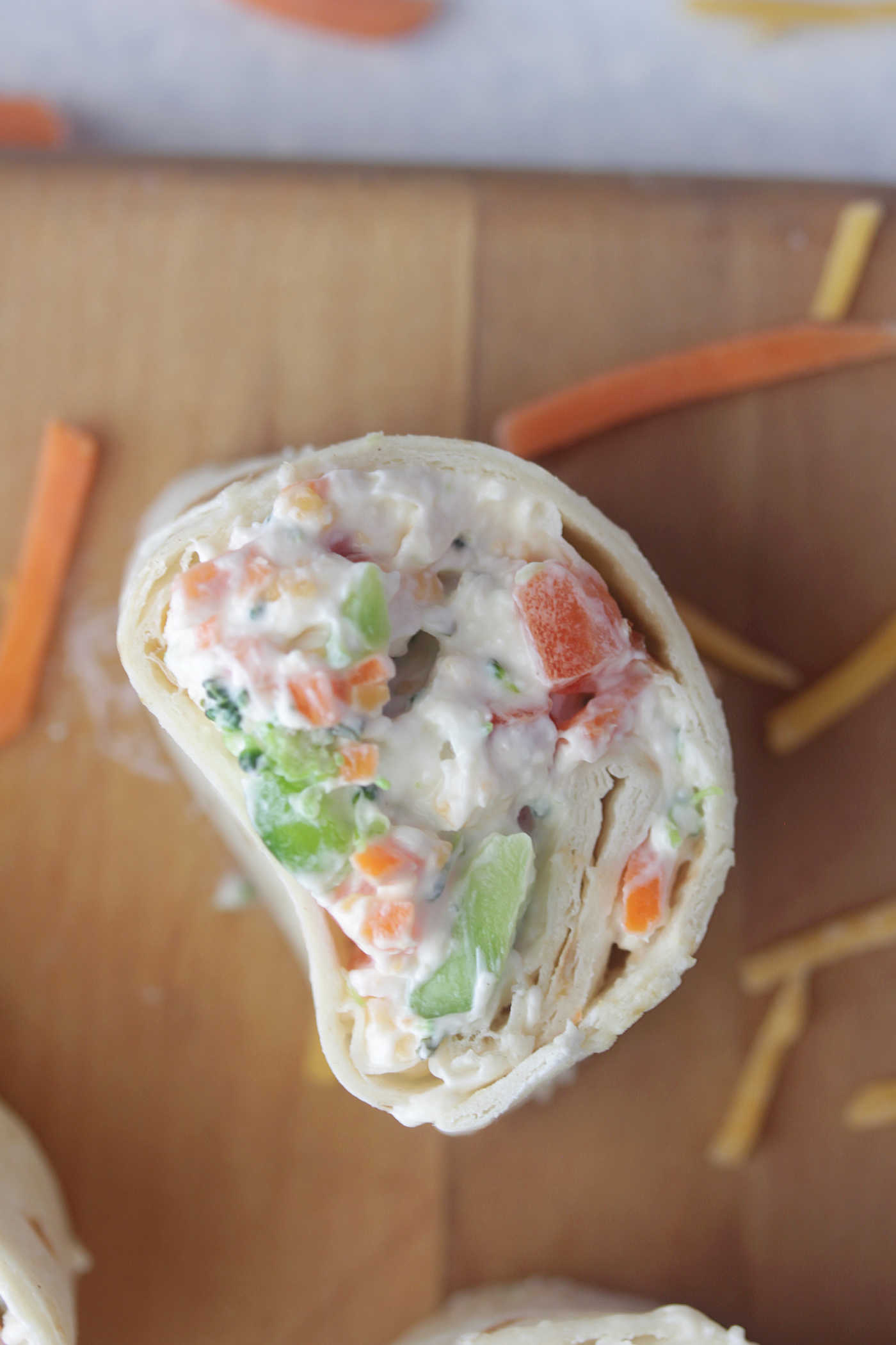Overhead photo of a veggie roll up on a wooden board surrounded by shredded carrots and cheddar cheese